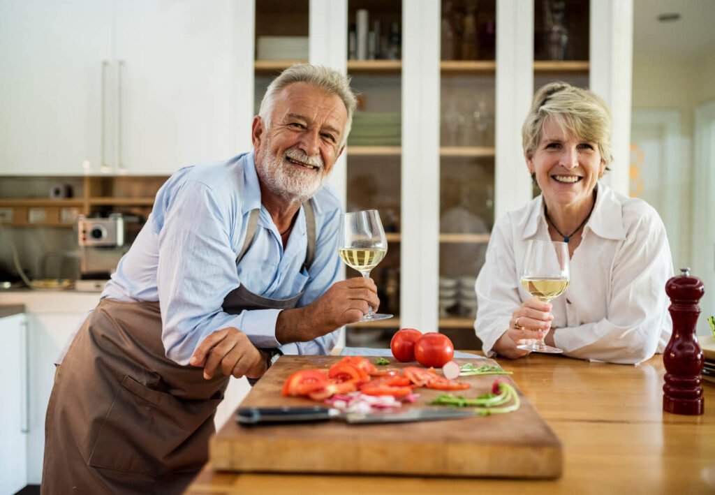 Casal idoso sorrindo na cozinha com taças de vinho, celebrando o resultado da prótese protocolo com carga imediata em São José dos Campos.